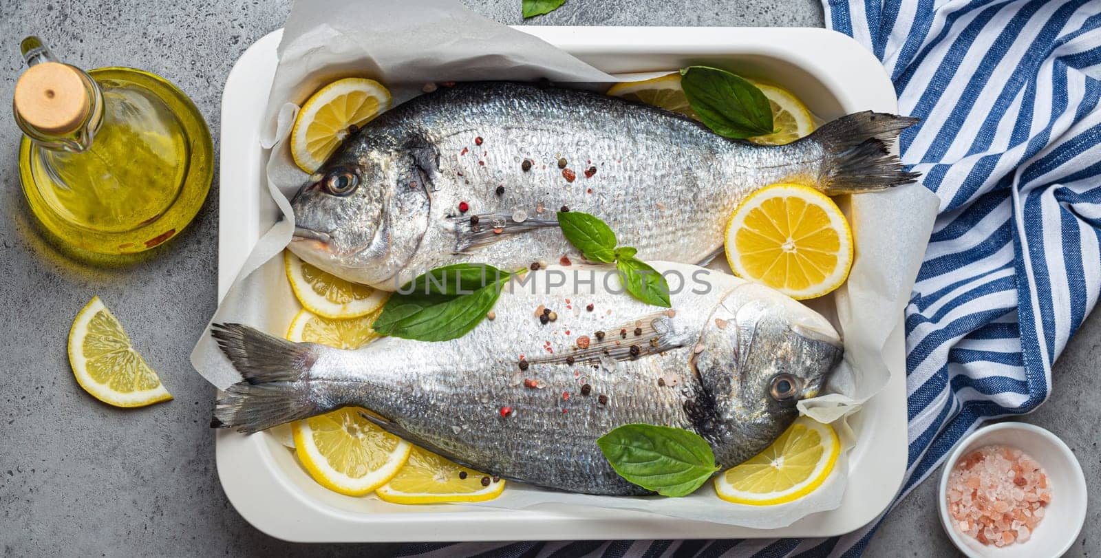 Raw fish dorado in casserole dish with ingredients lemon, fresh basil, bottle of olive oil on wooden cutting board with knife on rustic stone background top view, cooking healthy fish dorado concept