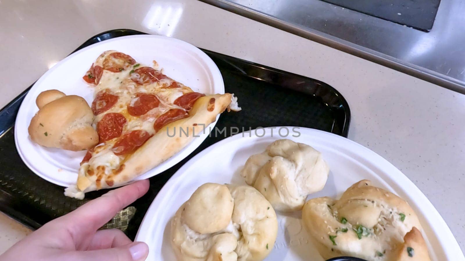 Close-up of a freshly baked pizza slice on a paper plate, placed on a black tray in a pizzeria. The background shows more pizzas being prepared, creating a warm and inviting atmosphere.