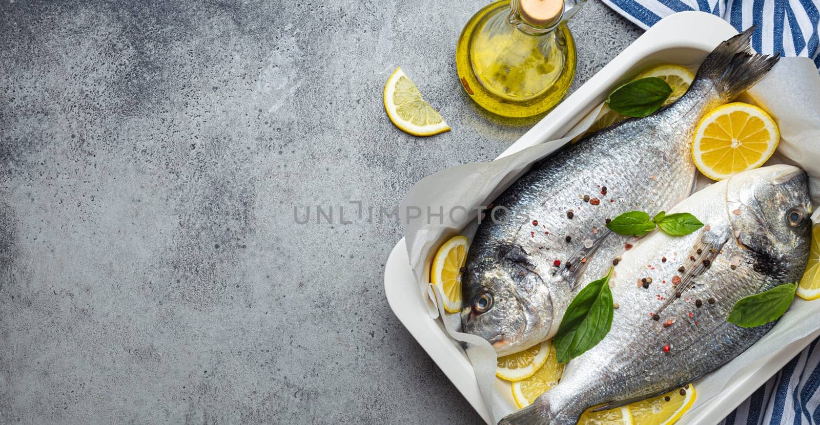 Raw fish dorado in casserole dish with ingredients lemon, fresh basil, bottle of olive oil on wooden cutting board on rustic stone background top view, cooking healthy fish dorado. Copy space