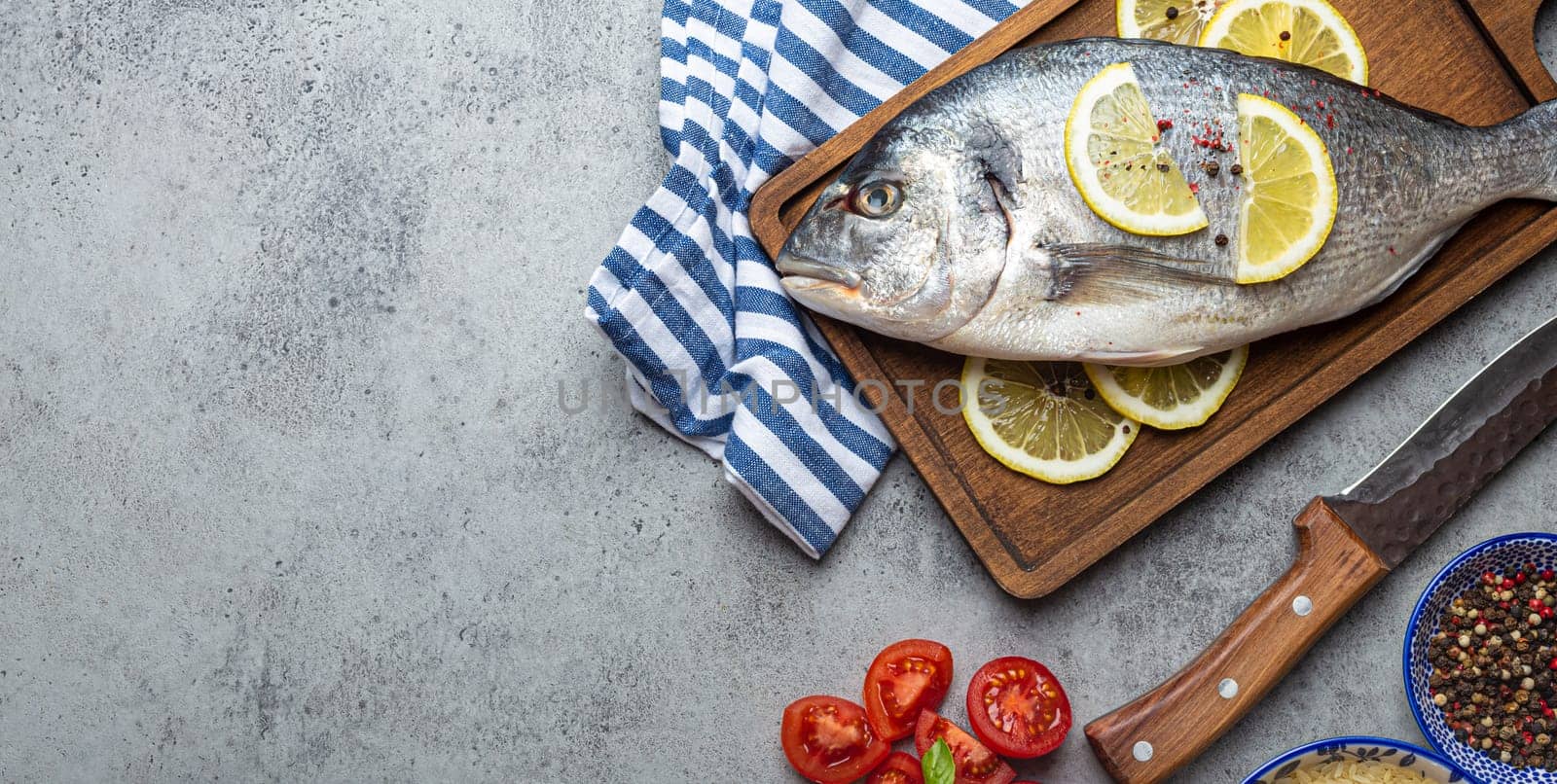 Raw fish dorado with ingredients lemon, fresh basil, cut cherry tomatoes, uncooked rice on wooden cutting board with knife on rustic stone background top view, cooking healthy fish dorado. Copy space