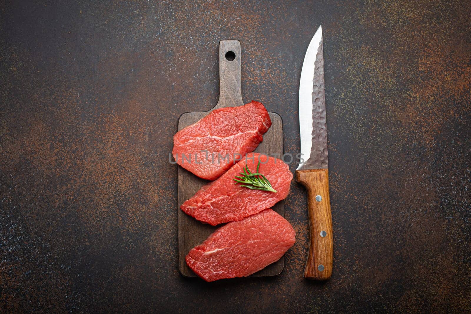 Three raw uncooked meat beef top sirloin steaks on wooden cutting board with rosemary on wooden cutting board with knife on dark rustic background from above, preparing meal with meat