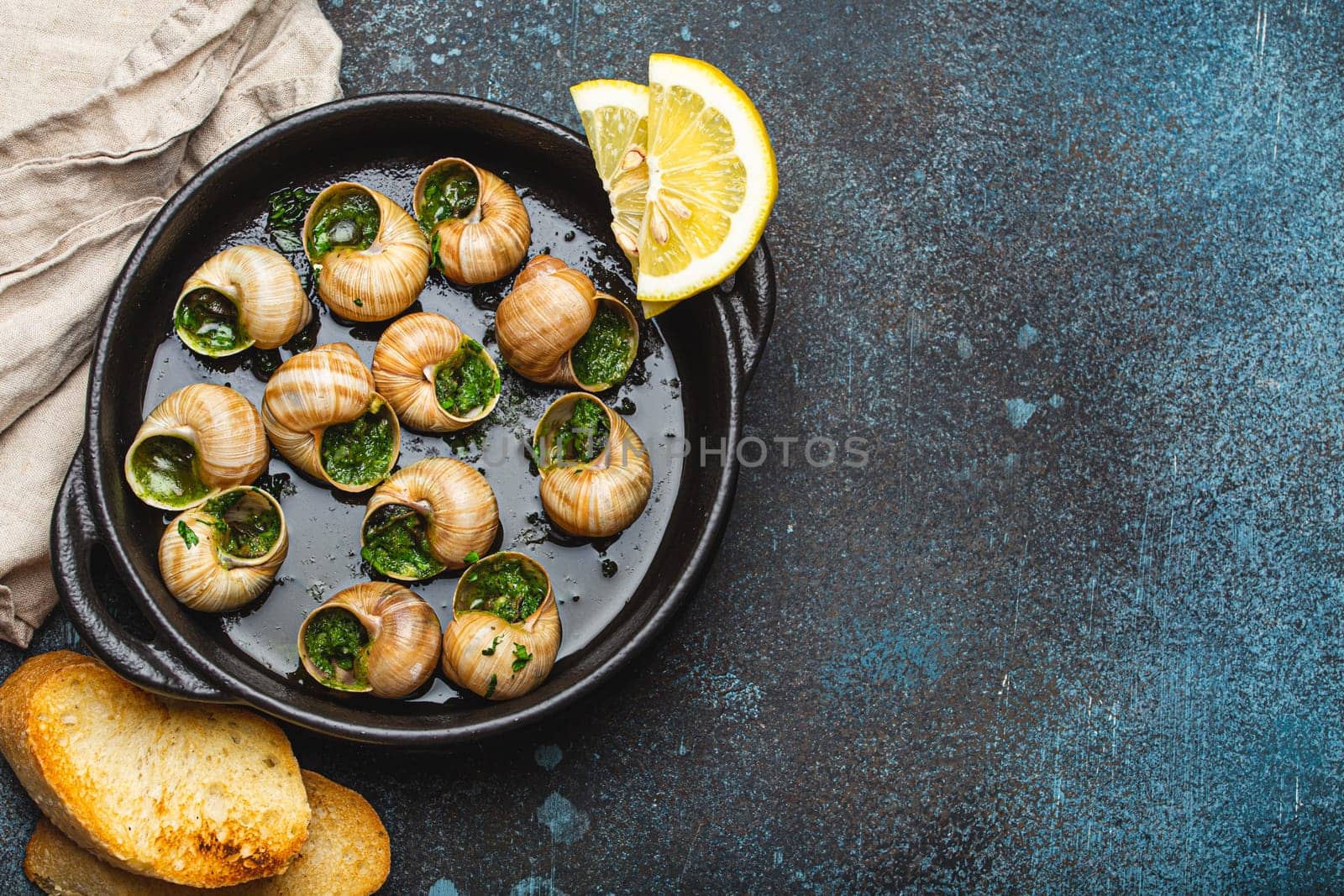 Escargots de Bourgogne Snails with Garlic Butter and Parsley in black cast iron pan with Lemon and Toasted Baguette Slices on rustic stone background top view, traditional French Delicacy, copy space.