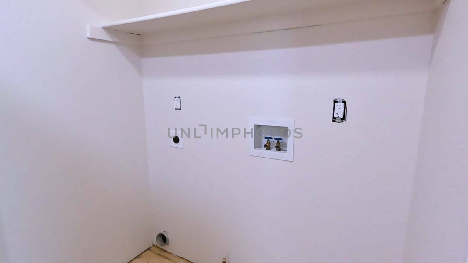 A utility room in the middle of renovation, showcasing exposed plumbing and electrical outlets. The space is prepped for the installation of a washer and dryer, with freshly painted walls and a built-in shelf.