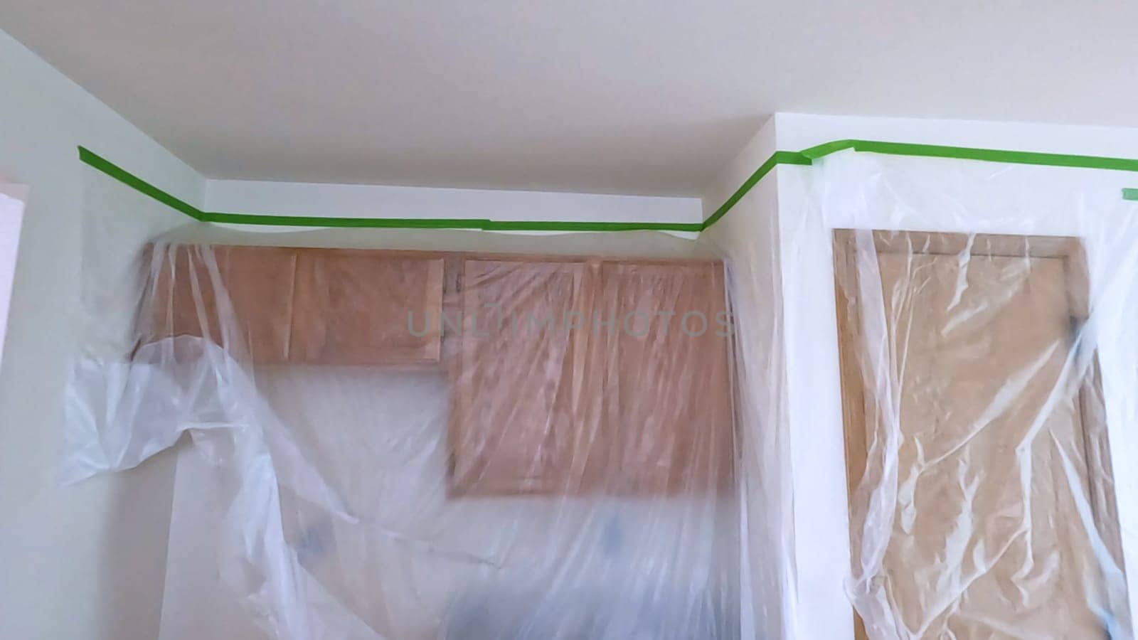 A kitchen undergoing renovation, featuring cabinets covered with plastic sheeting and masking tape along the walls and ceiling. The space is prepared for painting or further construction work.