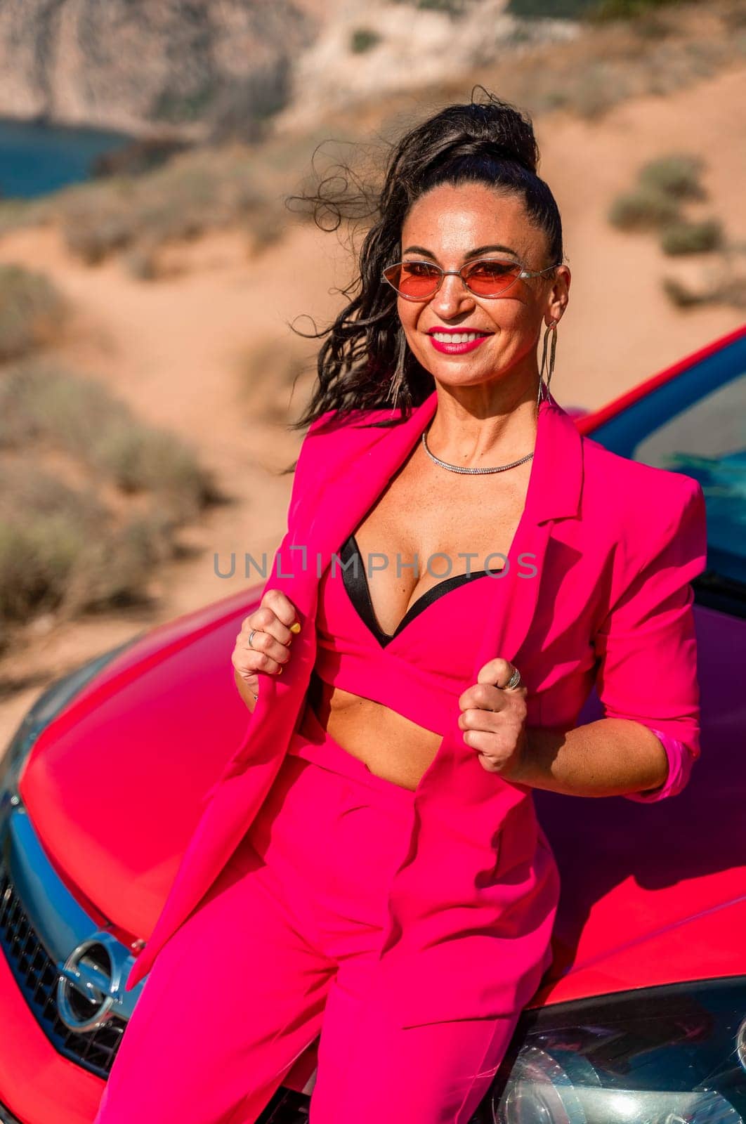 A woman in a pink suit is sitting on a red car. She is smiling and posing for the camera. The scene has a casual and relaxed vibe, with the woman enjoying her time outdoors. by Matiunina