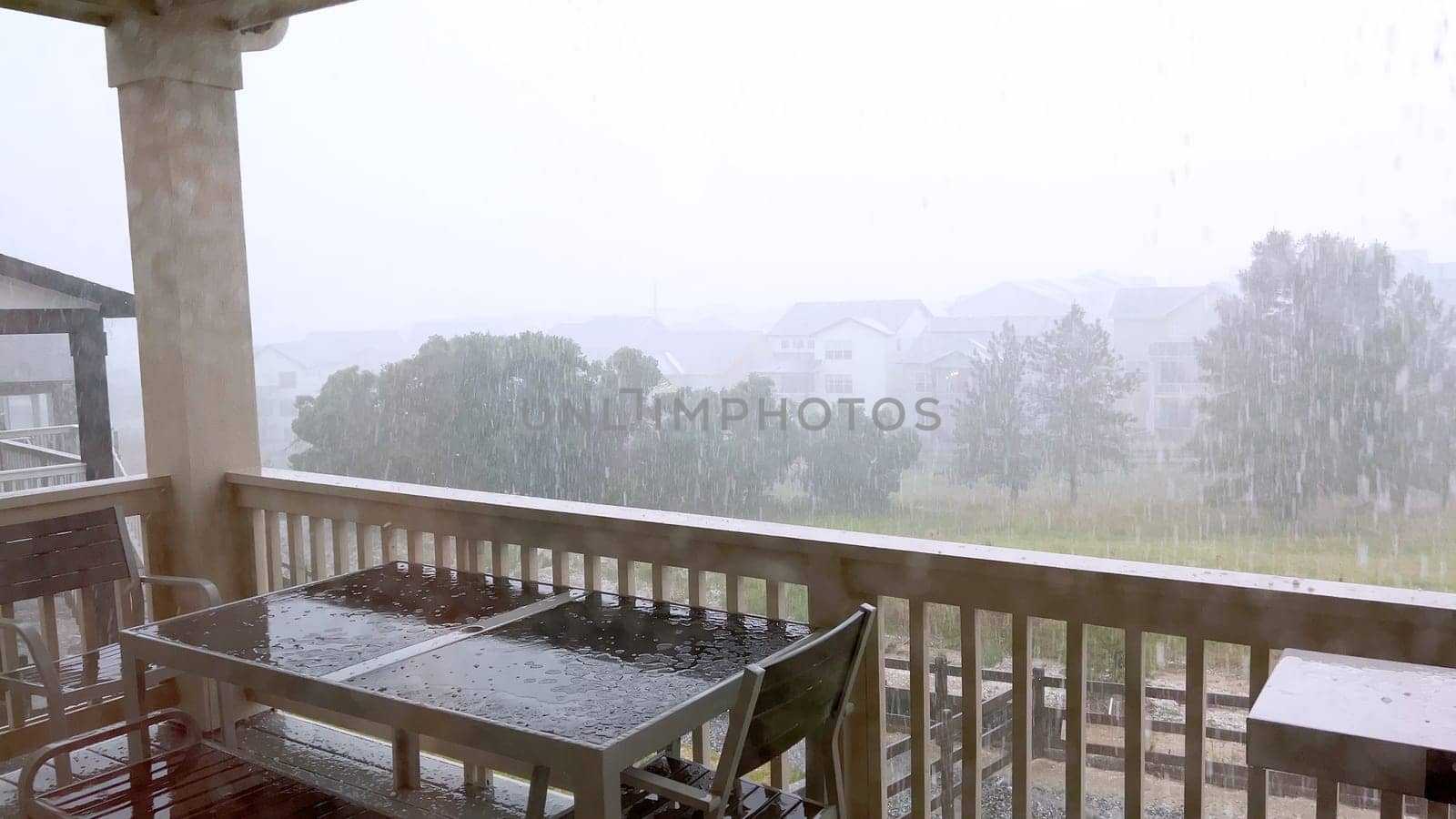 Image capturing a hail storm on a wooden deck, highlighting the impact of hail on patio furniture and the deck surface. Hailstones are visible scattered across the wet deck, with patio furniture in the background.