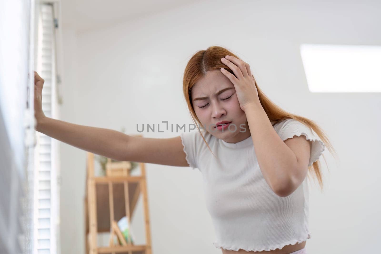 A young woman experiencing a severe headache, holding her head in pain while standing in a bright, modern room.