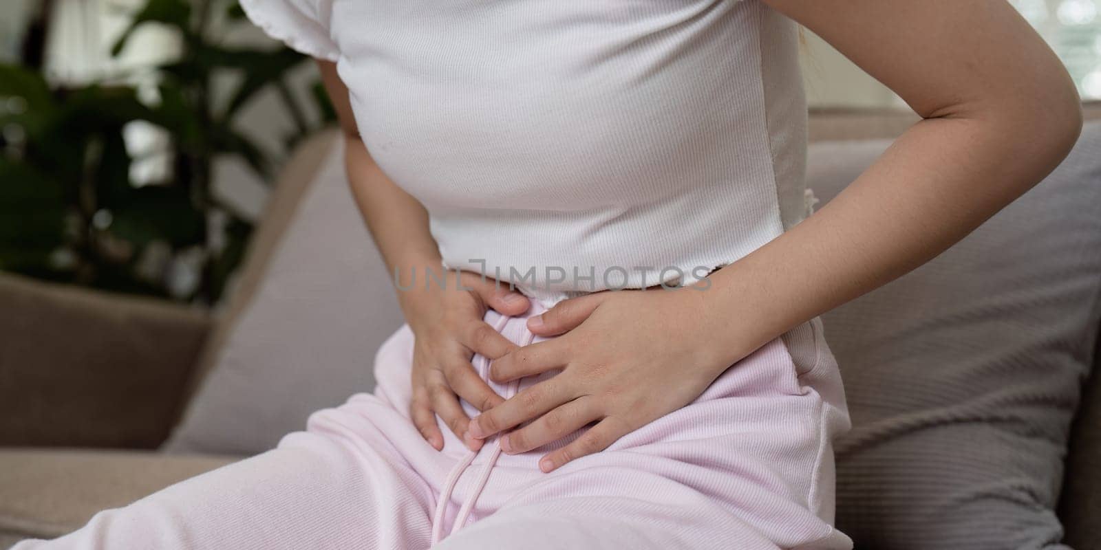 Woman Experiencing Stomach Pain While Sitting on a Couch in a Home Setting, Clutching Her Abdomen in Discomfort by nateemee