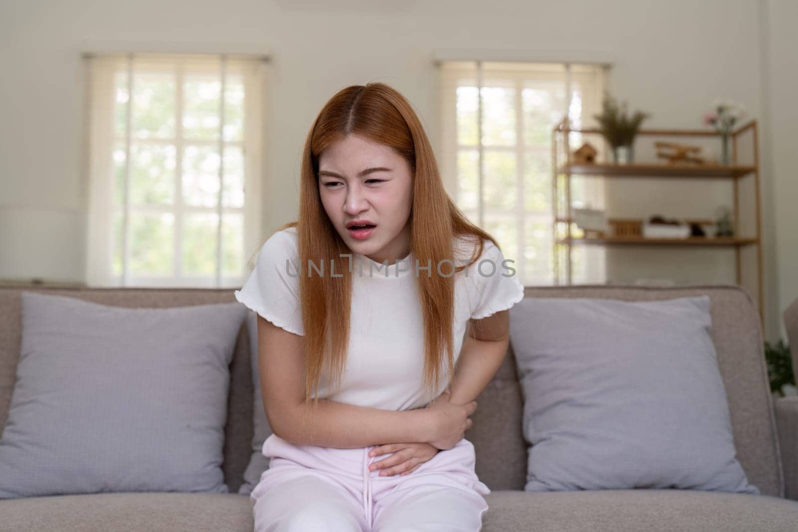 A young woman holding her stomach in pain while sitting on a couch in a bright, modern living room.