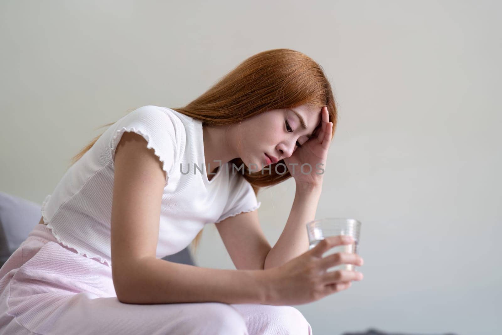 A young woman with a headache, holding a glass of water, sitting in a minimalist home environment, conveying discomfort and pain.