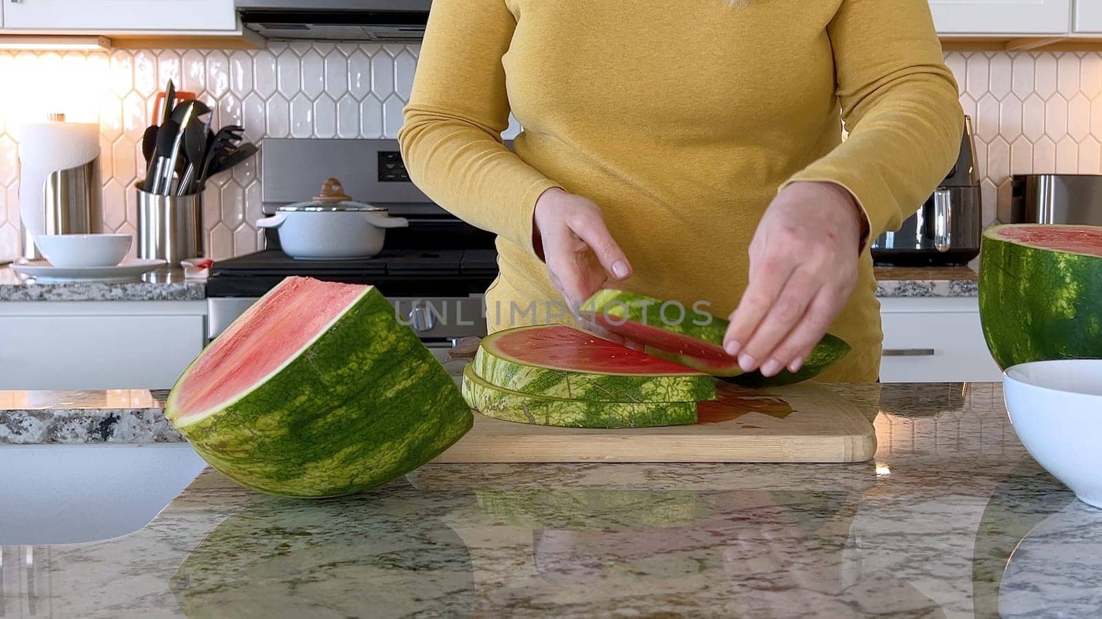 A woman in a yellow sweater slices a fresh watermelon on a cutting board in a modern kitchen. The kitchen features white cabinetry, a hexagonal tile backsplash, and various cooking utensils on the countertop.