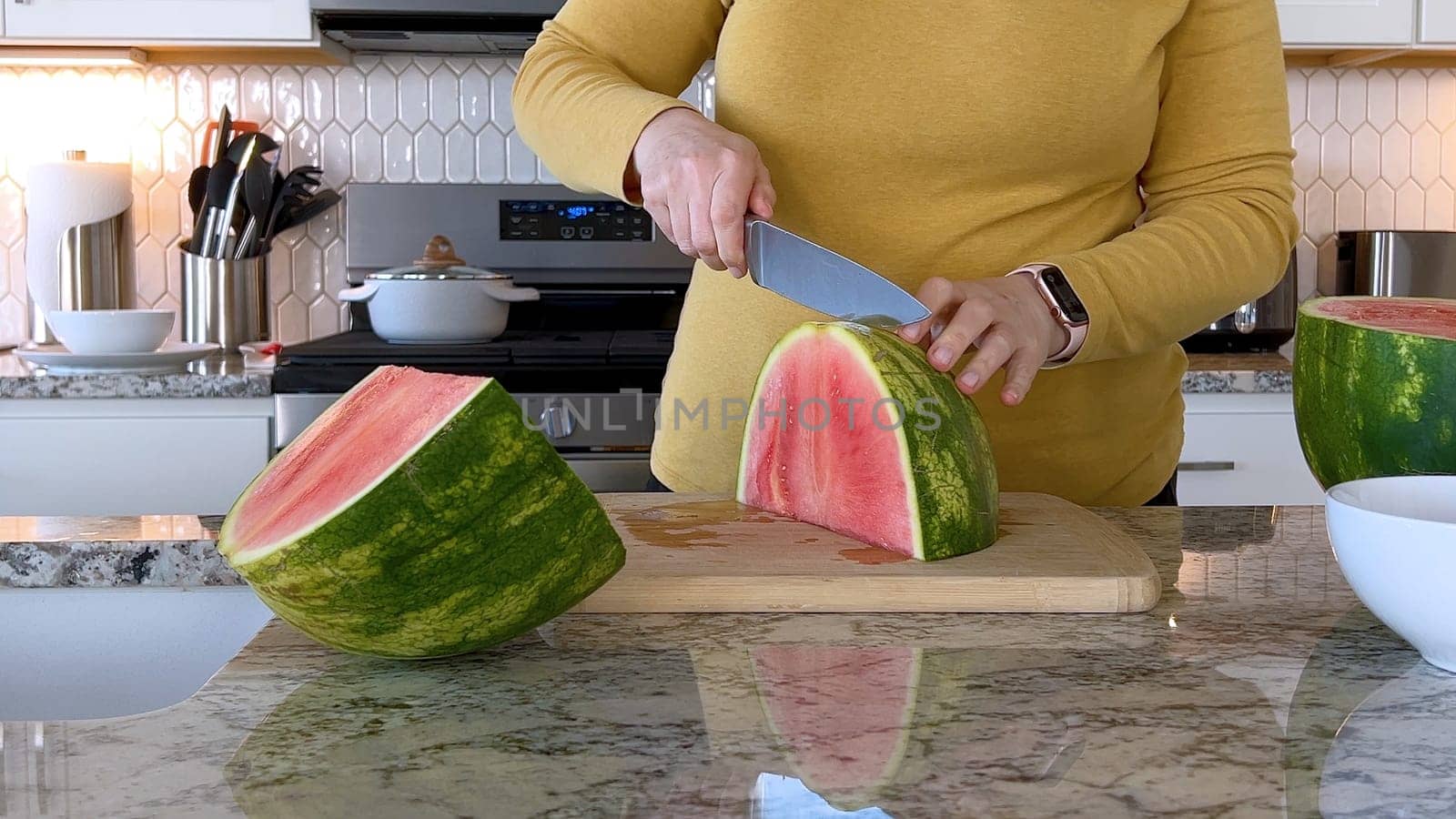 A woman in a yellow sweater slices a fresh watermelon on a cutting board in a modern kitchen. The kitchen features white cabinetry, a hexagonal tile backsplash, and various cooking utensils on the countertop.