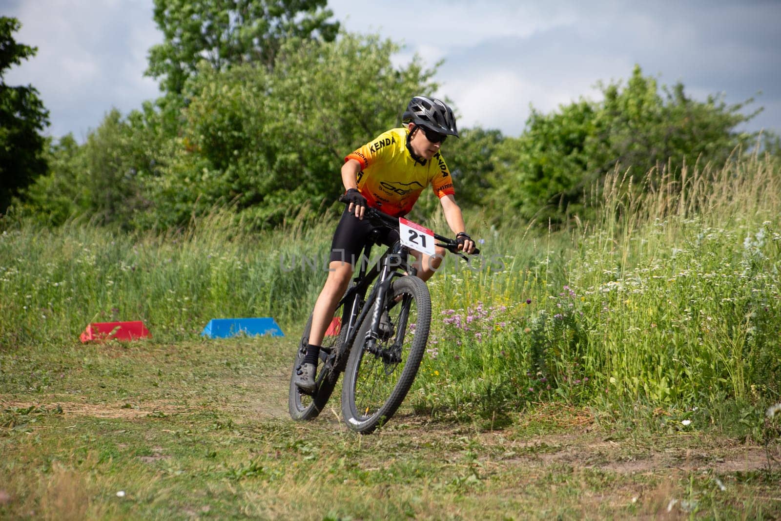 Kursk, Russia, June 15, 2024: Young cyclist at amateur competition rides bicycle over rough terrain against backdrop of tall grass on sunny day