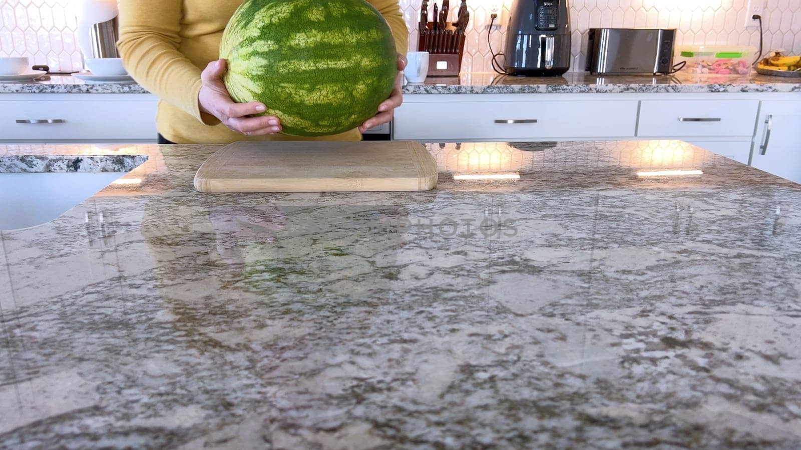 A woman in a yellow sweater slices a fresh watermelon on a cutting board in a modern kitchen. The kitchen features white cabinetry, a hexagonal tile backsplash, and various cooking utensils on the countertop.