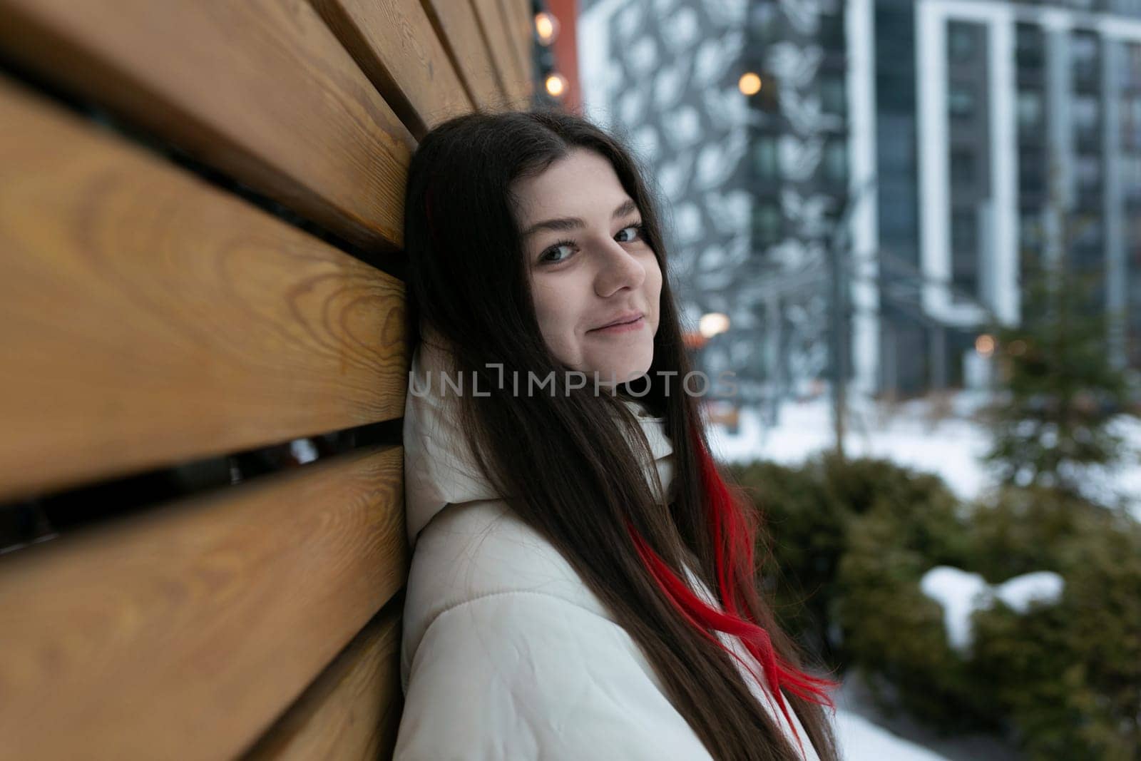 A woman leaning against a wooden bench covered in snow. The snow-covered ground and bench contrast with the dark clothing worn by the woman. She appears to be looking off into the distance.