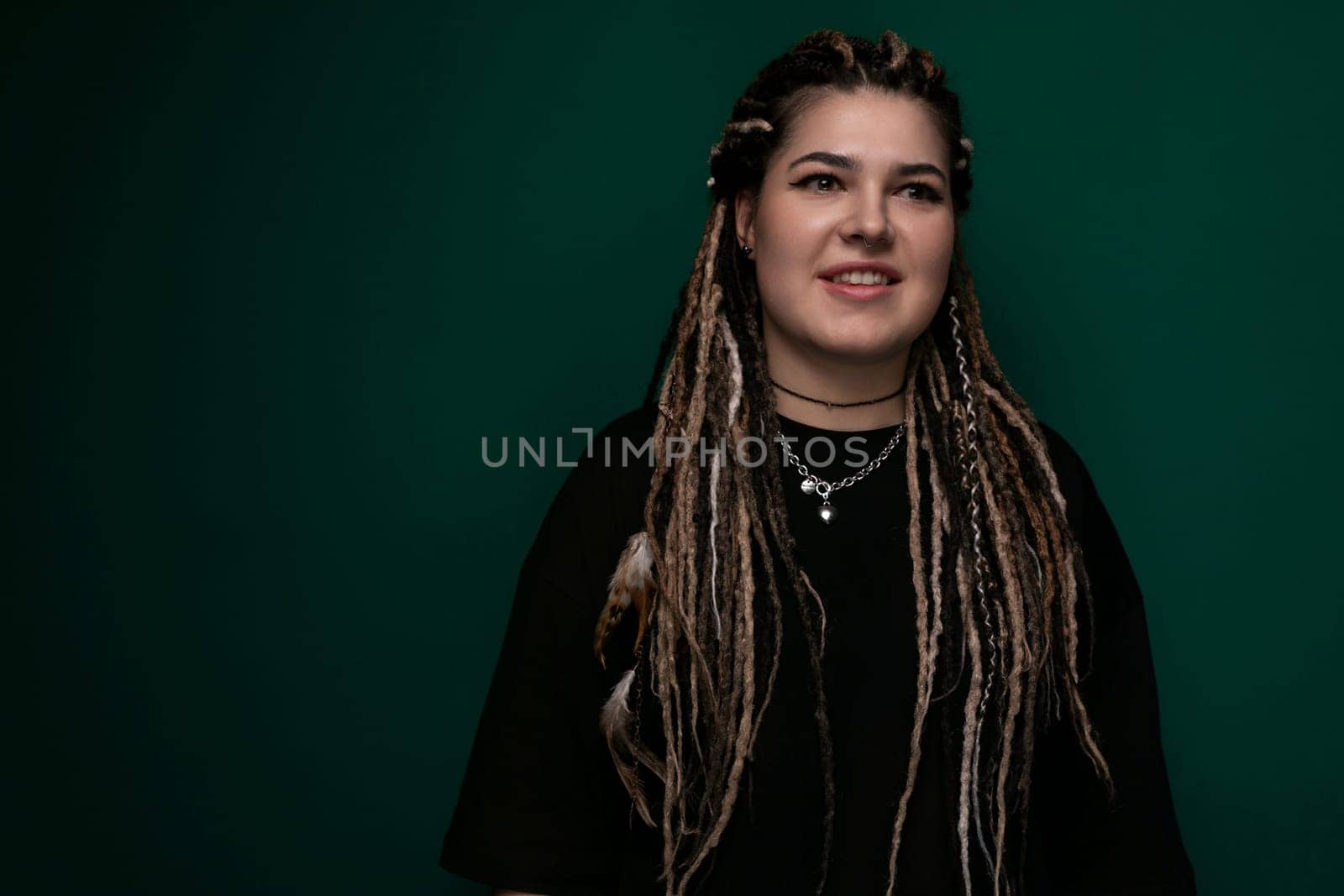 A woman with dreadlocks is standing in front of a bright green background. She is looking directly at the camera with a neutral expression. The womans hair is styled in long, thin, twisted dreadlocks, and she is wearing casual clothing.