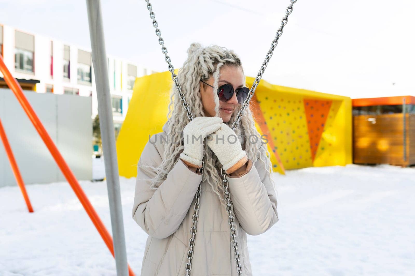 A woman wearing a white coat and sunglasses is sitting on a swing. The swing is in motion, with the woman looking towards the camera.