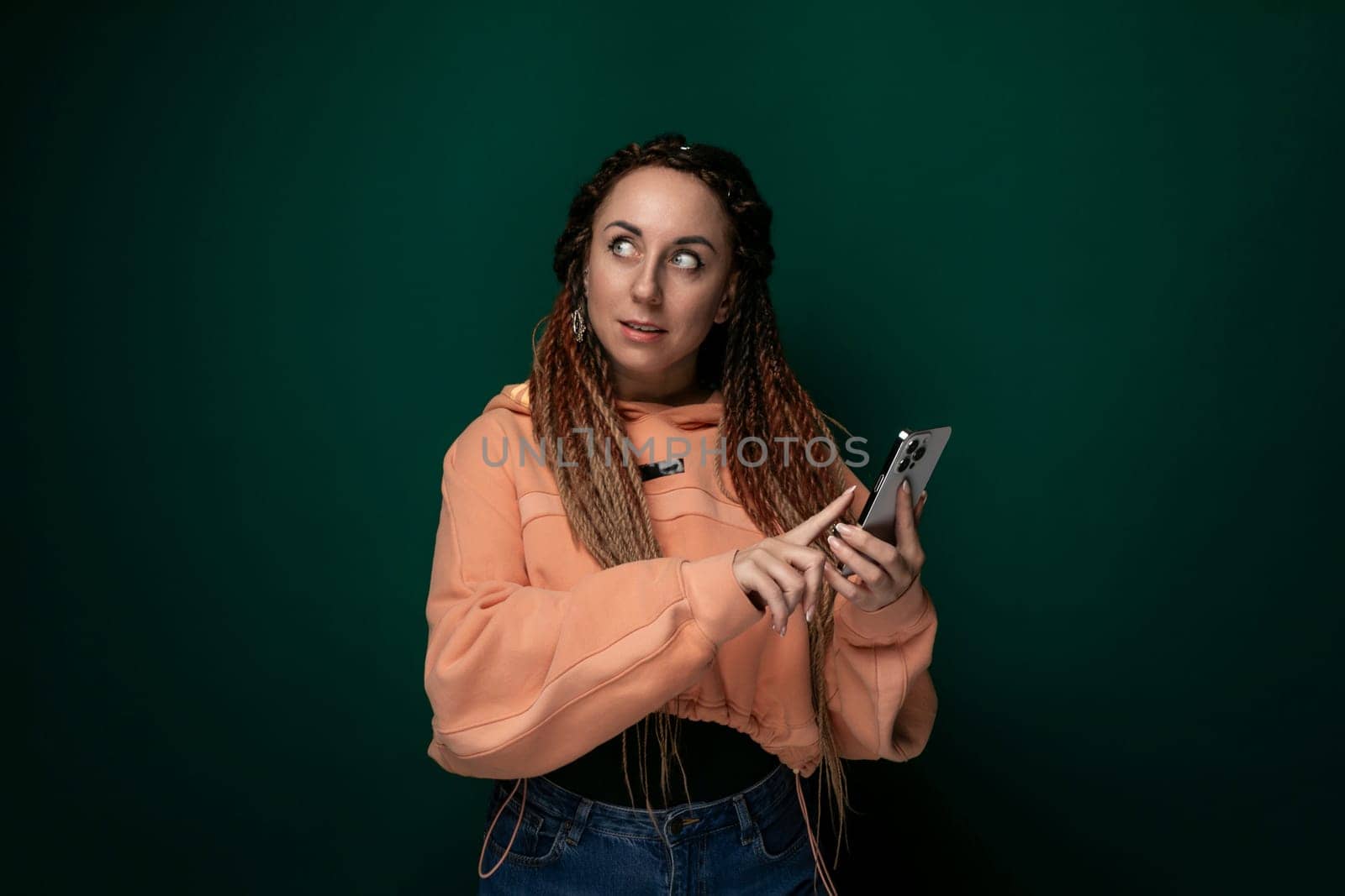 A woman with dreadlocks is shown holding a cell phone in her hand. She appears to be using the phone and looking at the screen. Her hair is styled in thick, rope-like dreadlocks. The background is nondescript.