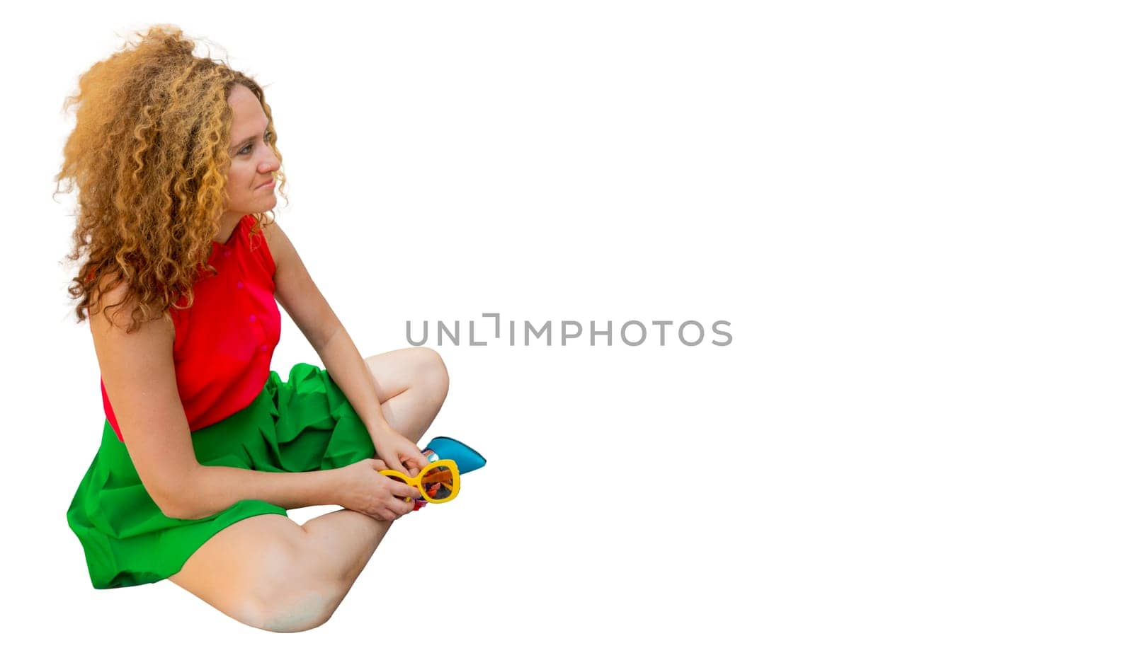 Fashionable woman in bright clothes, with long blond curly hair, is sitting in the lotus position on the street basking in the sun and resting against the background of a modern business center.