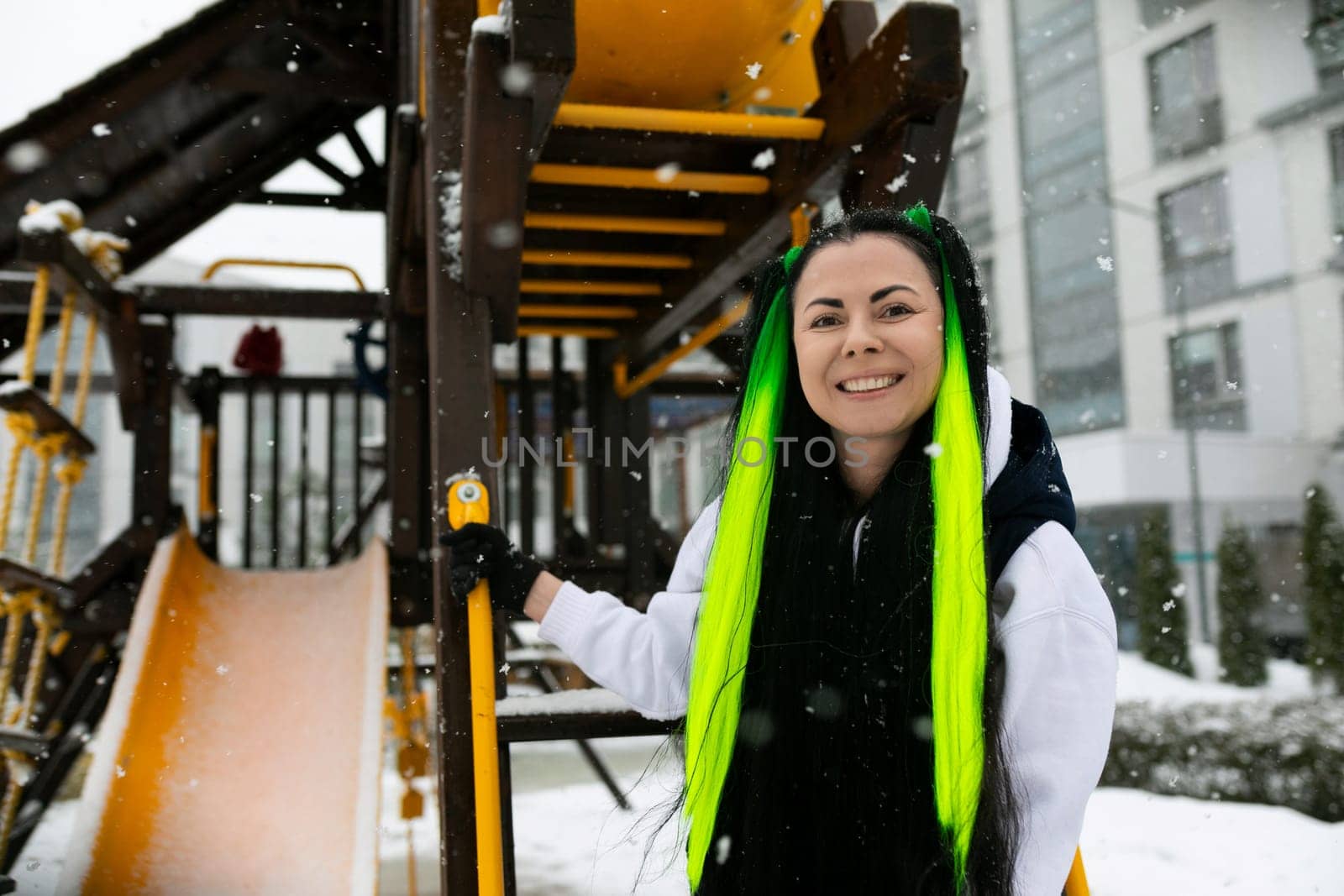 A woman with vibrant green hair stands confidently in front of a colorful playground, looking towards the camera. She is surrounded by swings, slides, and other playground equipment.