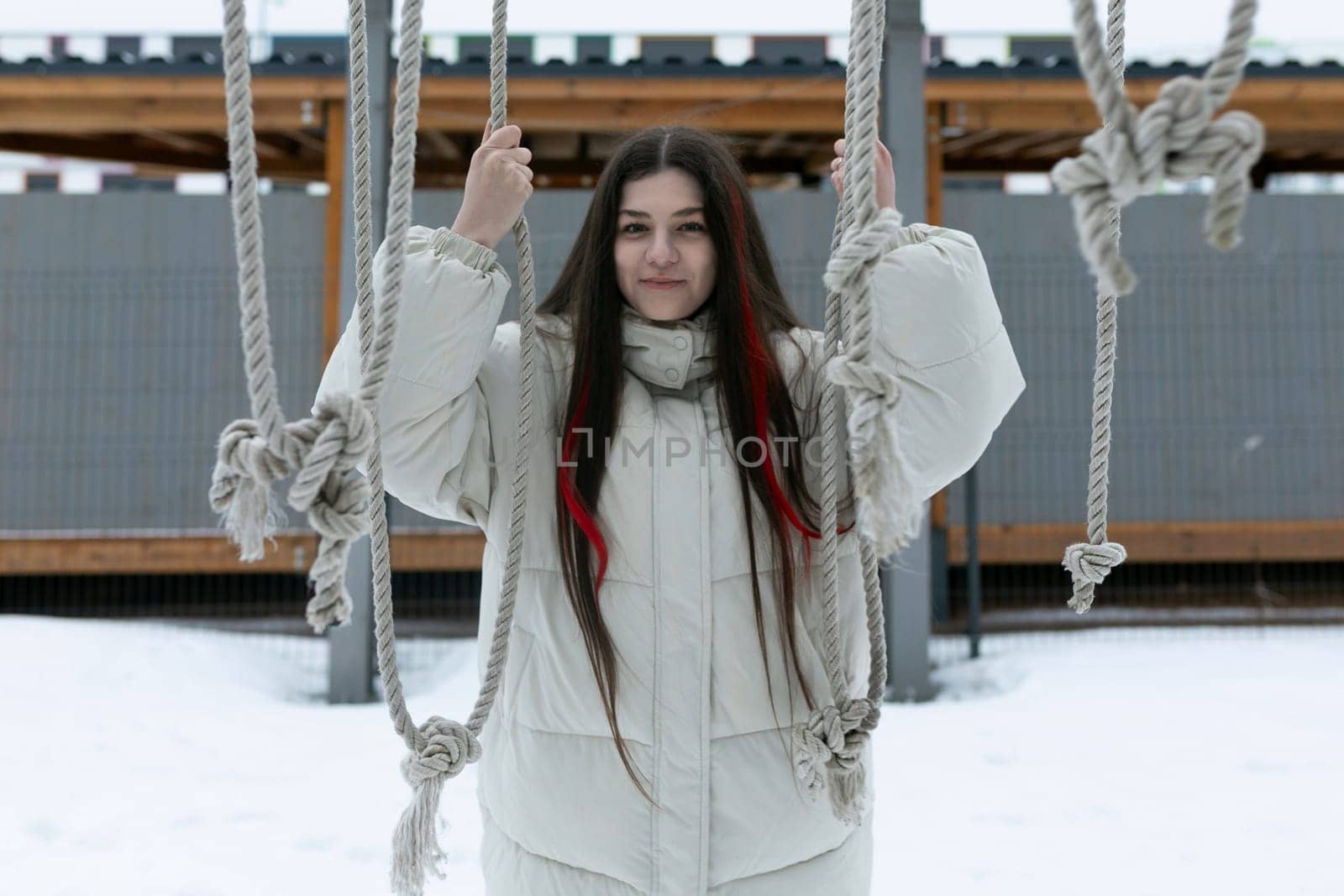 A woman in warm clothing stands on a swing covered in snow, holding onto the chains for balance. The snow-covered ground and trees in the background indicate a winter setting.