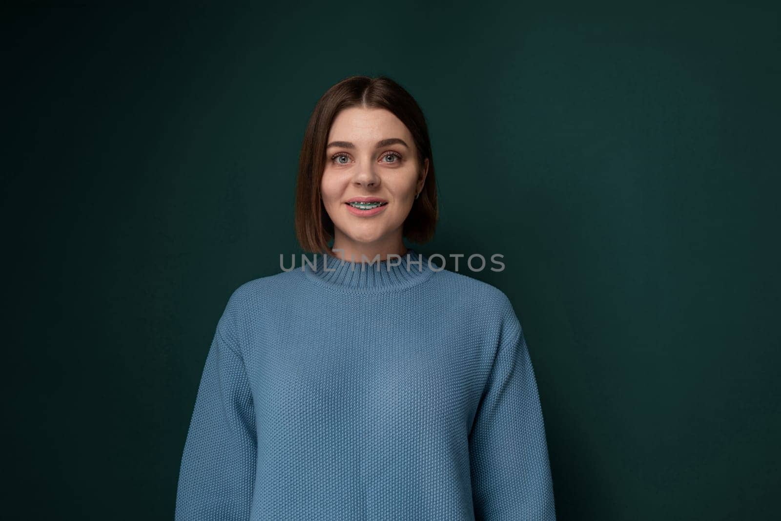 A woman standing in front of a plain green wall, looking directly at the camera. She appears relaxed and confident, with a neutral expression. The background is unadorned and focuses the viewers attention on the woman.