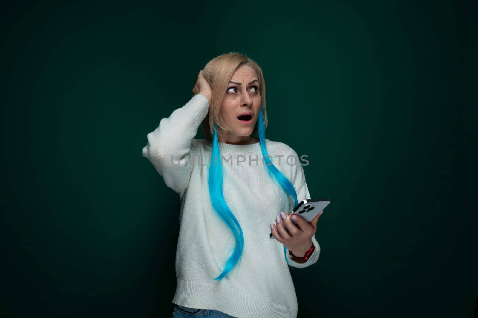 A woman with vibrant blue hair is holding a modern cell phone in her hand. She appears focused on the devices screen as she stands against a neutral background.