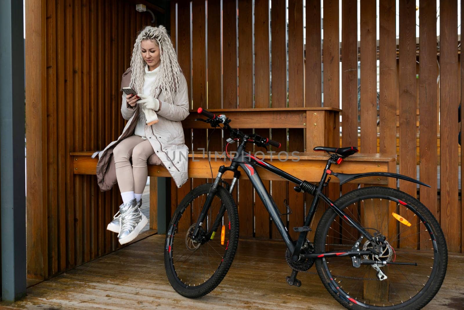 A woman is sitting on a bench next to a parked bicycle. She appears relaxed and is looking around. The bike is leaning against the bench, and there are other benches and trees in the background.