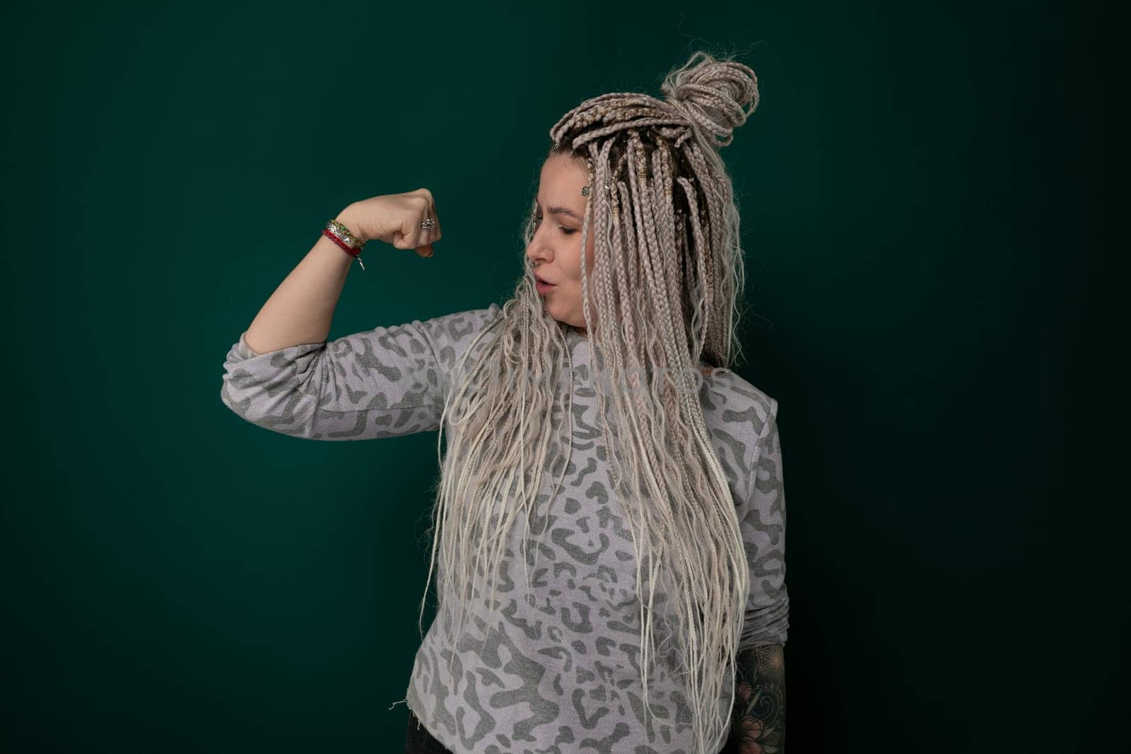 A woman with long blonde dreadlocks is standing in front of a green wall. She appears confident and stylish in her unique hairstyle, contrasting with the vibrant background.