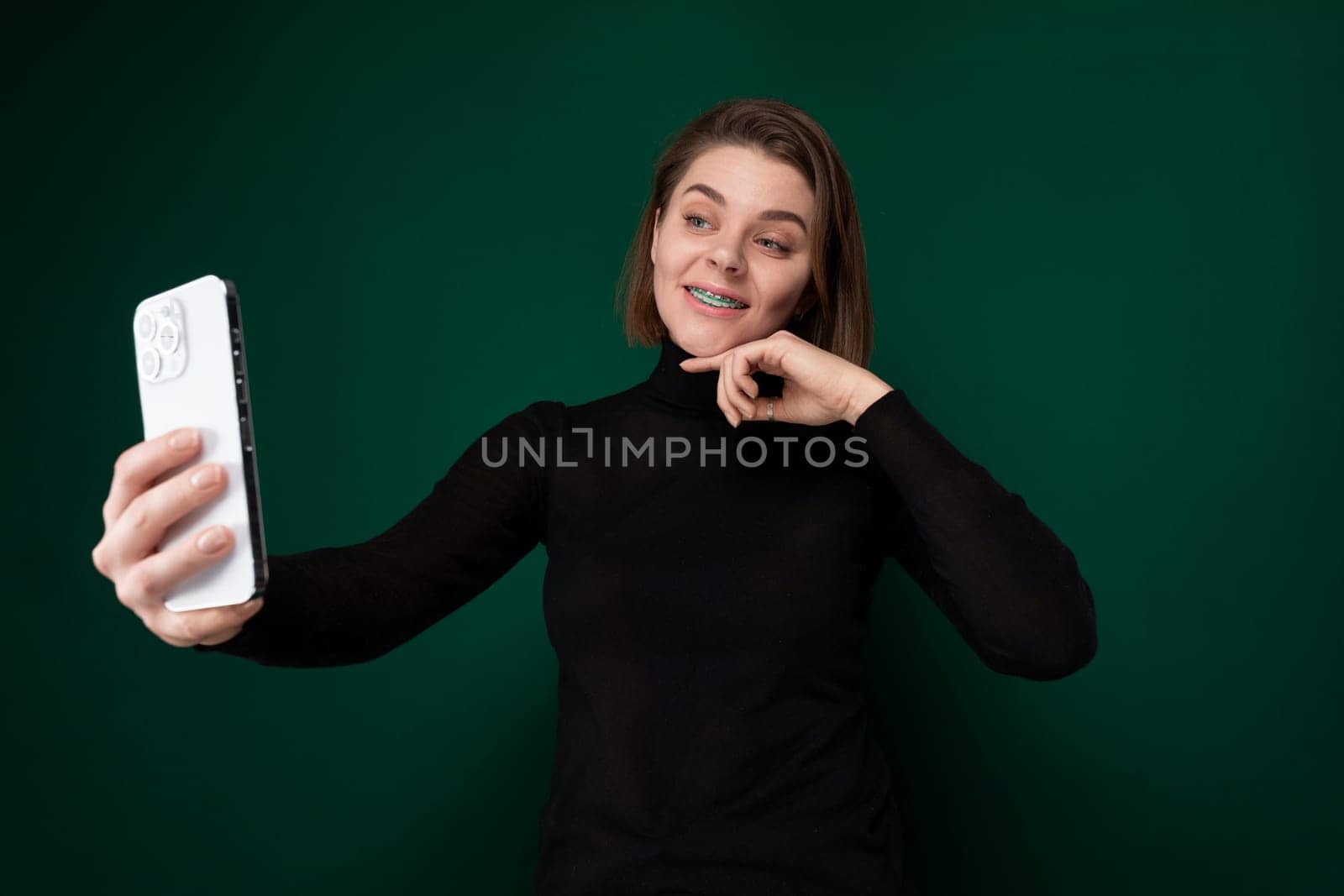 A woman is standing outdoors, holding her cell phone at arms length, and smiling as she takes a selfie.