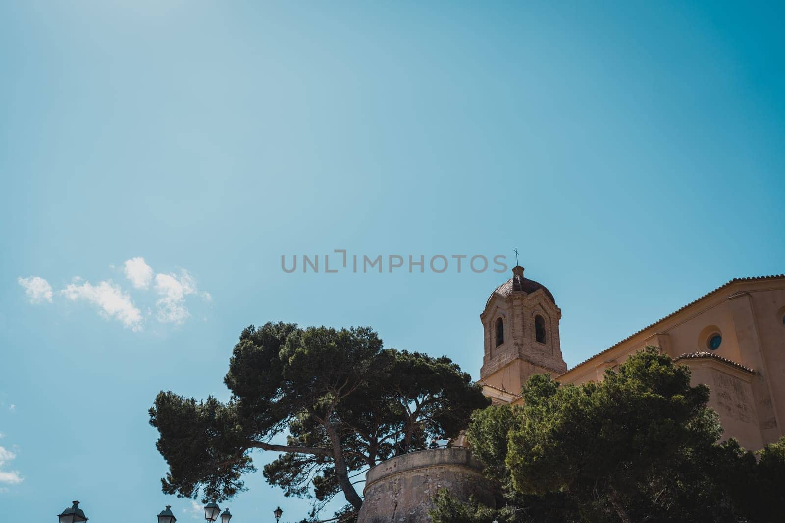 The tower of a historic church surrounded by trees against a clear blue sky. Cullera Castle, Spain