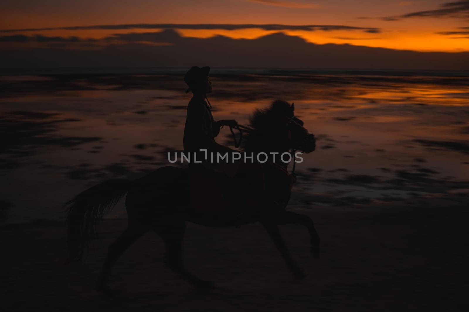 Horse unrecognized rider on the beach during sunset, captured in silhouette against a vibrant sky. The scene reflects on the wet sand, creating a picturesque, serene moment by the ocean.
