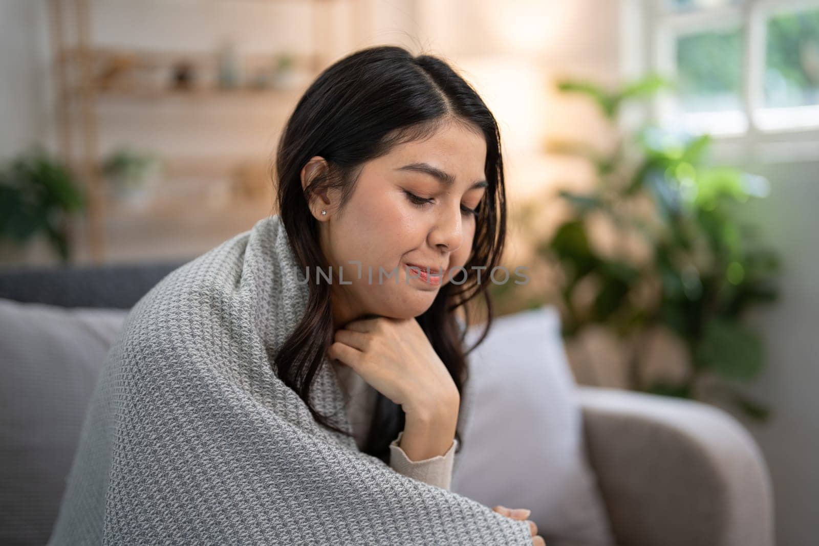 Young woman with pharyngitis feeling unwell, wrapped in a blanket while sitting on a sofa, demonstrating the concept of feeling sick and healthcare