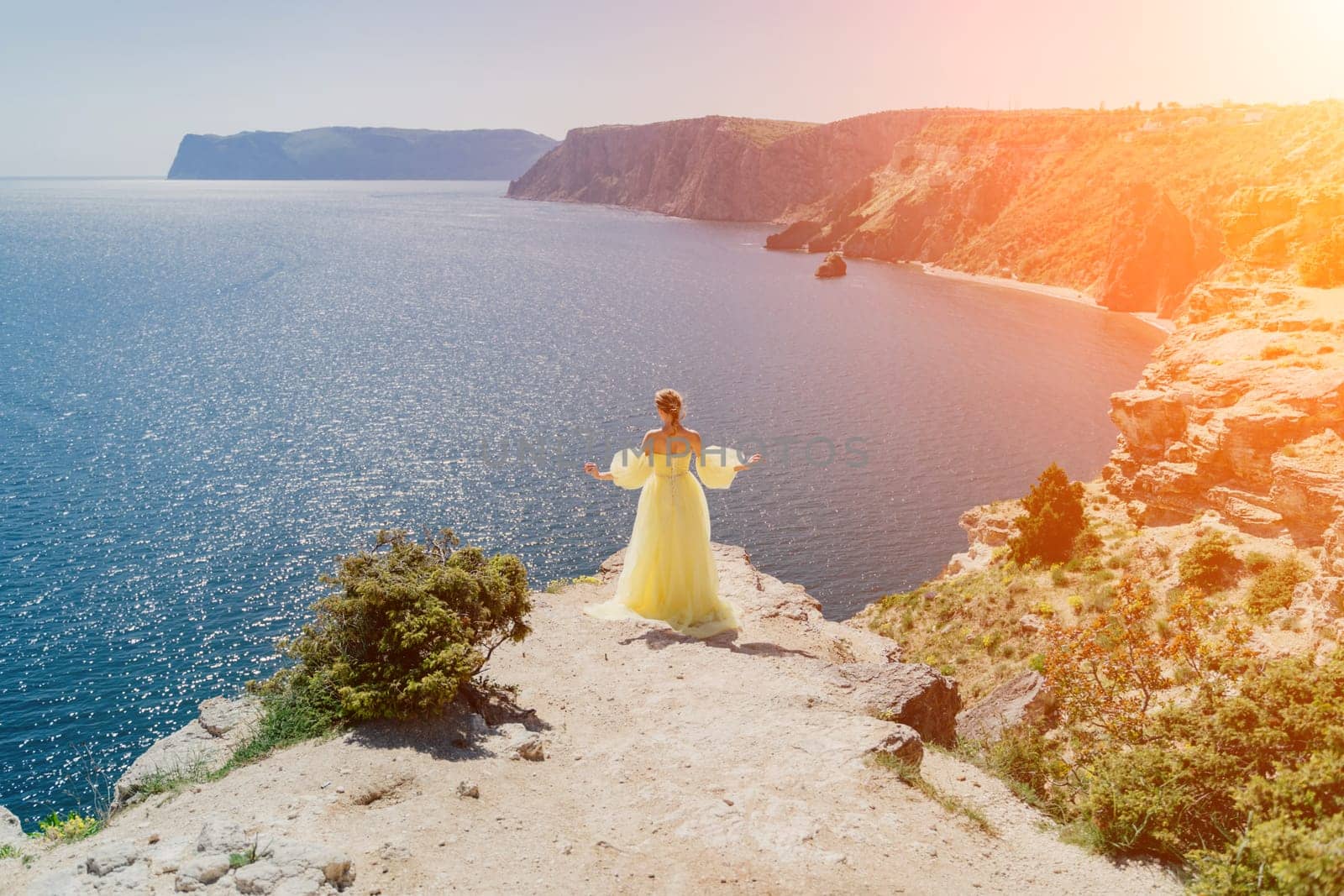 Woman yellow dress sea. Side view Young beautiful sensual woman in yellow long dress posing on a rock high above the sea at sunset. Girl in nature against the blue sky by Matiunina