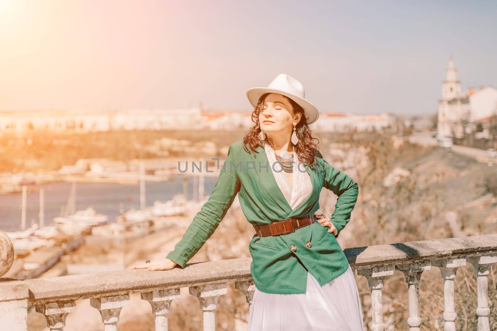 Woman walks around the city, lifestyle. Happy woman in a green jacket, white skirt and hat is sitting on a white fence with balusters overlooking the sea bay and the city. by Matiunina