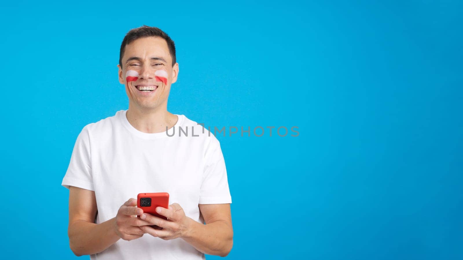 Polish supporter with the flag of Poland painted on his face, looking at his mobile phone smiling