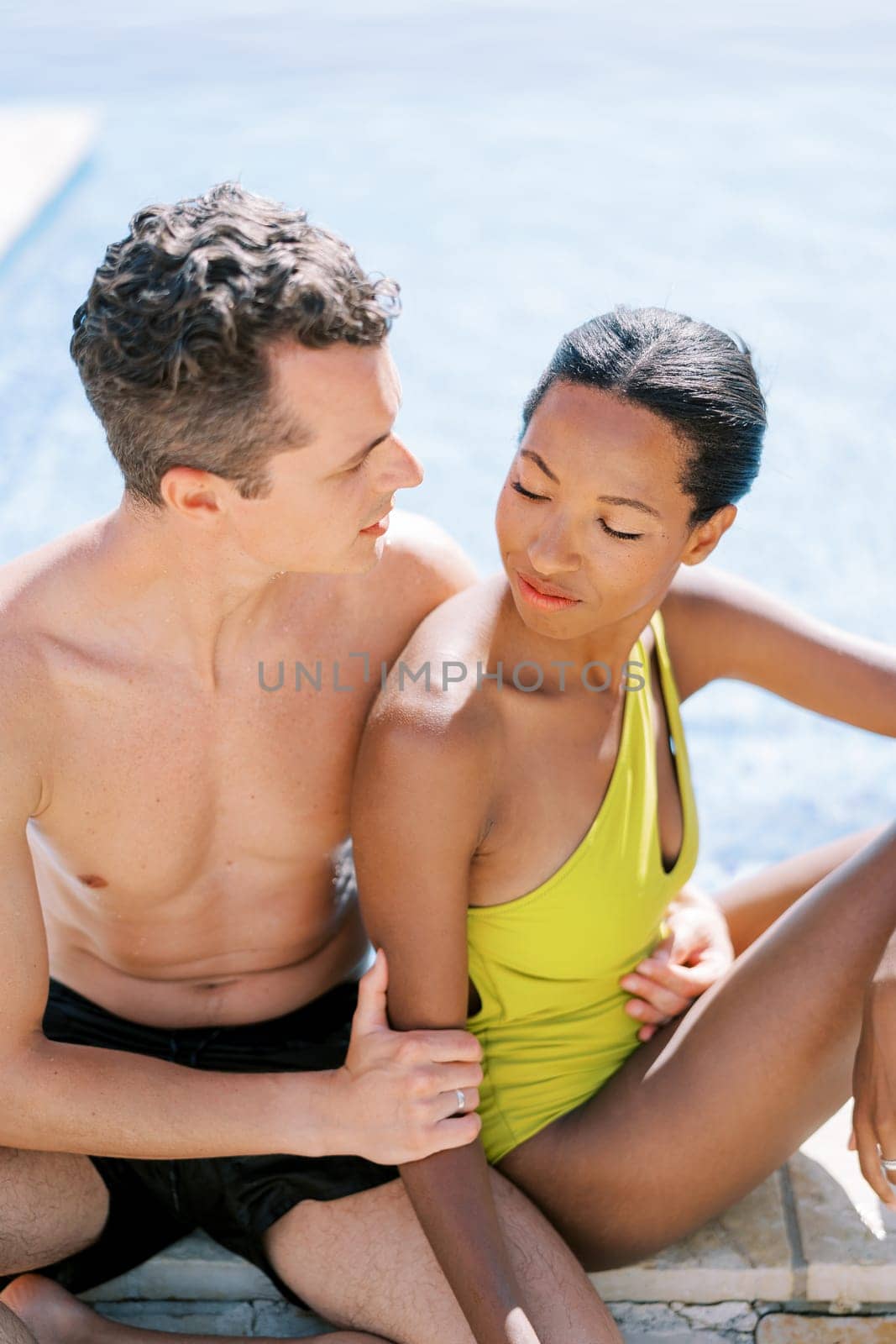 Man hugging woman waist from behind, looking at her, sitting on edge of pool. High quality photo