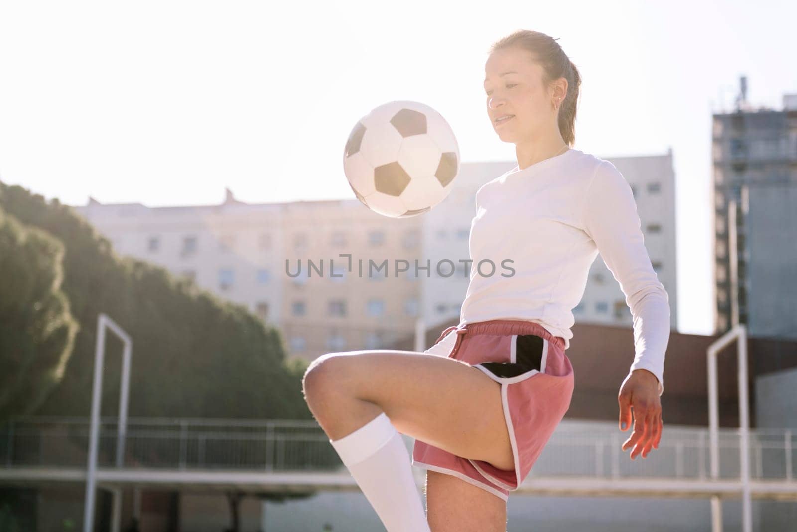 caucasian female soccer player kicking the ball with her knee on an urban football court, concept of sport and active lifestyle