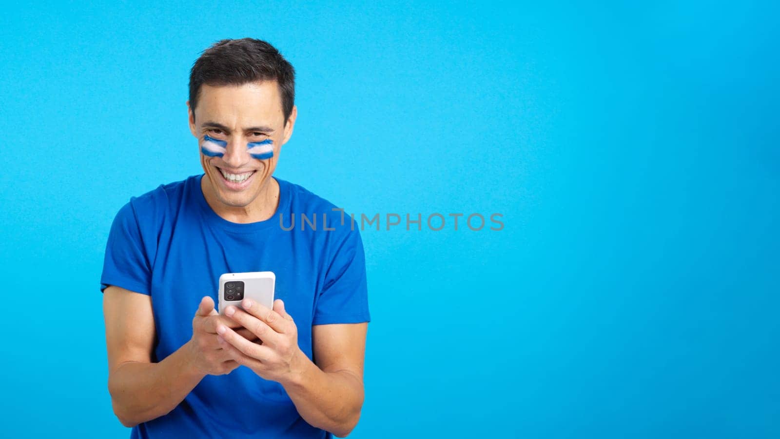 Salvadoran supporter with the flag of El Salvador painted on his face, looking at his mobile phone smiling