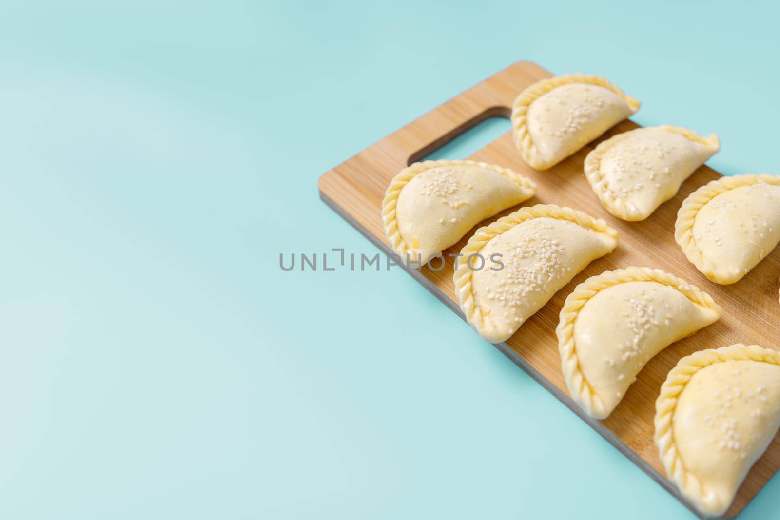 Raw samosas with sesame on a wooden on board on blue table ready for baking. Traditional Central Asian cuisine, frozen semi-finished products, copy space.