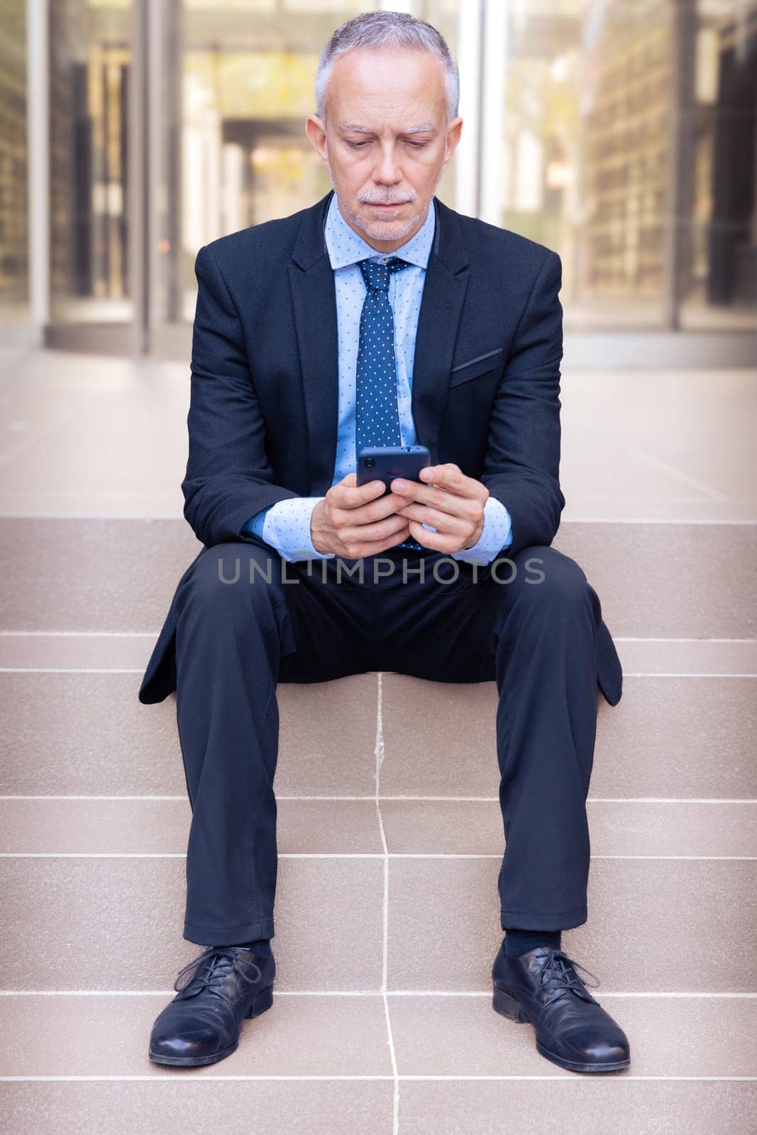 Satisfied with the results, the businessman walks down the street outside the office building, a mature boss holds a phone and coffee in his hands, writes messages and reads news online, using an app.