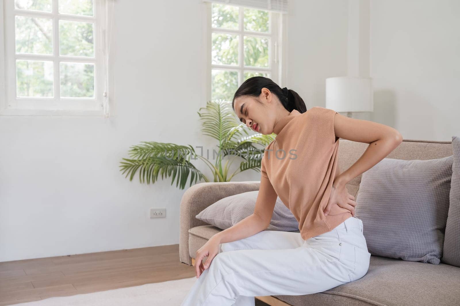 Young woman in casual clothing suffering from back pain, sitting on a couch in a bright, modern living room with green plants and natural light.