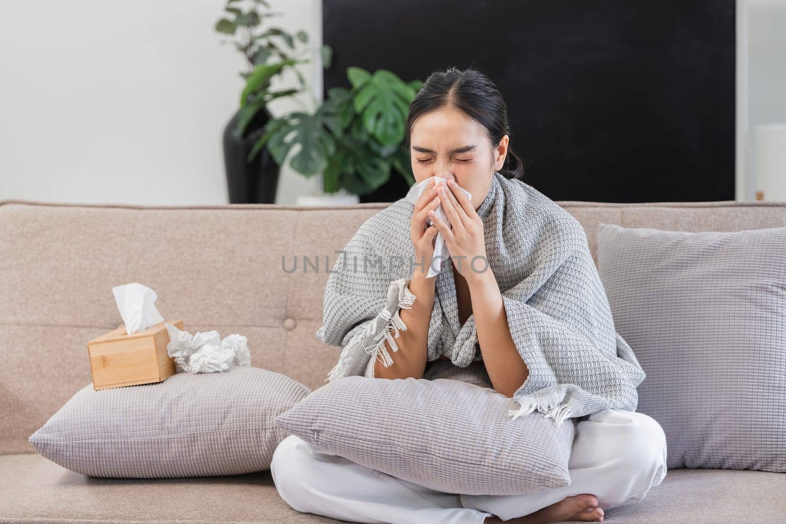 A woman with a fever sits on a sofa, wrapped in a blanket, holding a tissue, and looking unwell in a modern living room setting.