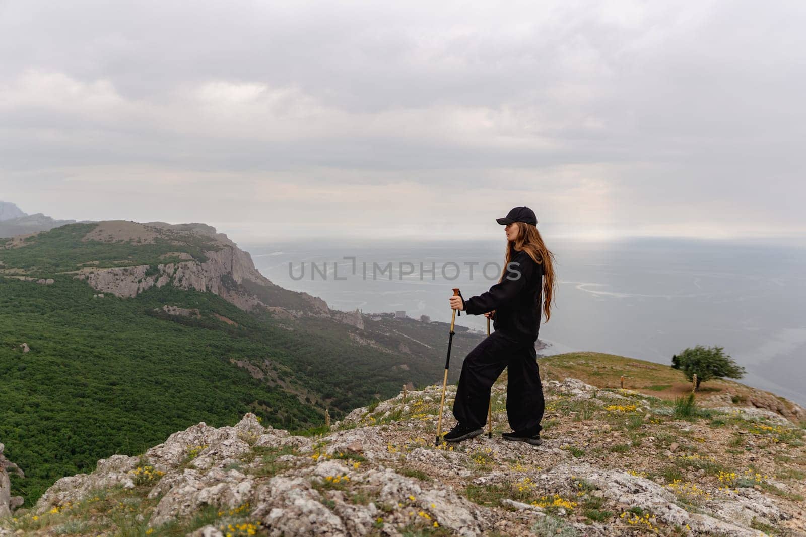 A woman stands on a mountain top, looking out over the ocean. The sky is cloudy, and the woman is wearing a black jacket