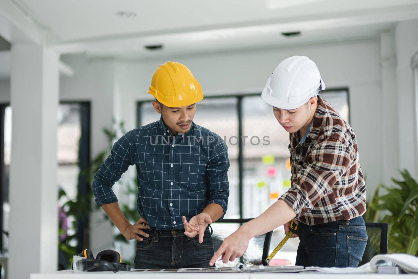 Two male engineers in a modern office, wearing safety helmets, reviewing blueprints, and discussing construction plans.