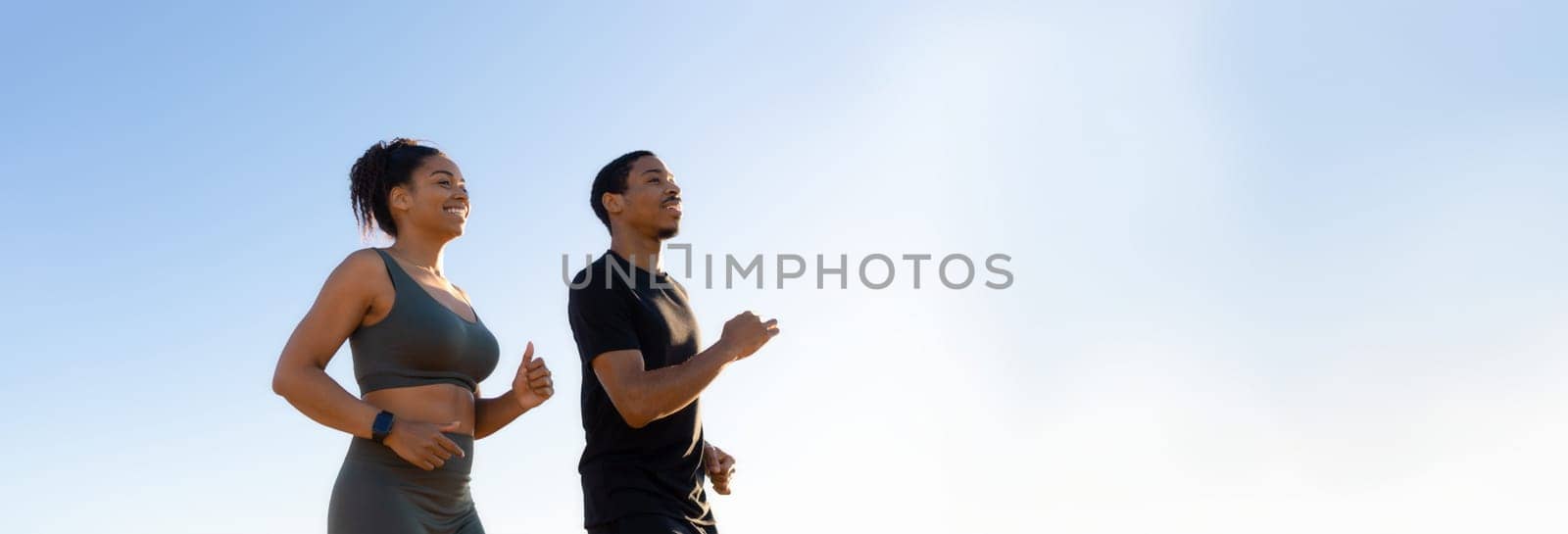Black woman and man are jogging side-by-side in front of a clear blue sky. They are both smiling and appear to be enjoying their workout, copy space