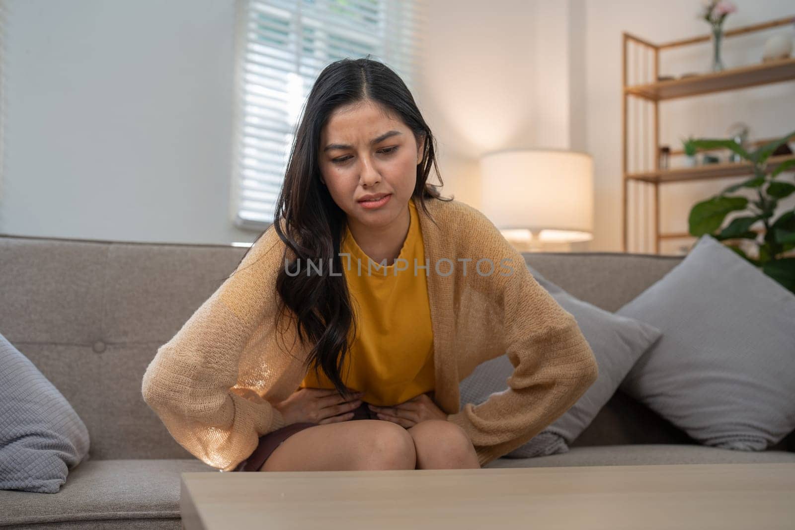 A young woman sitting on a couch appears to be in discomfort, clutching her stomach with a concerned expression