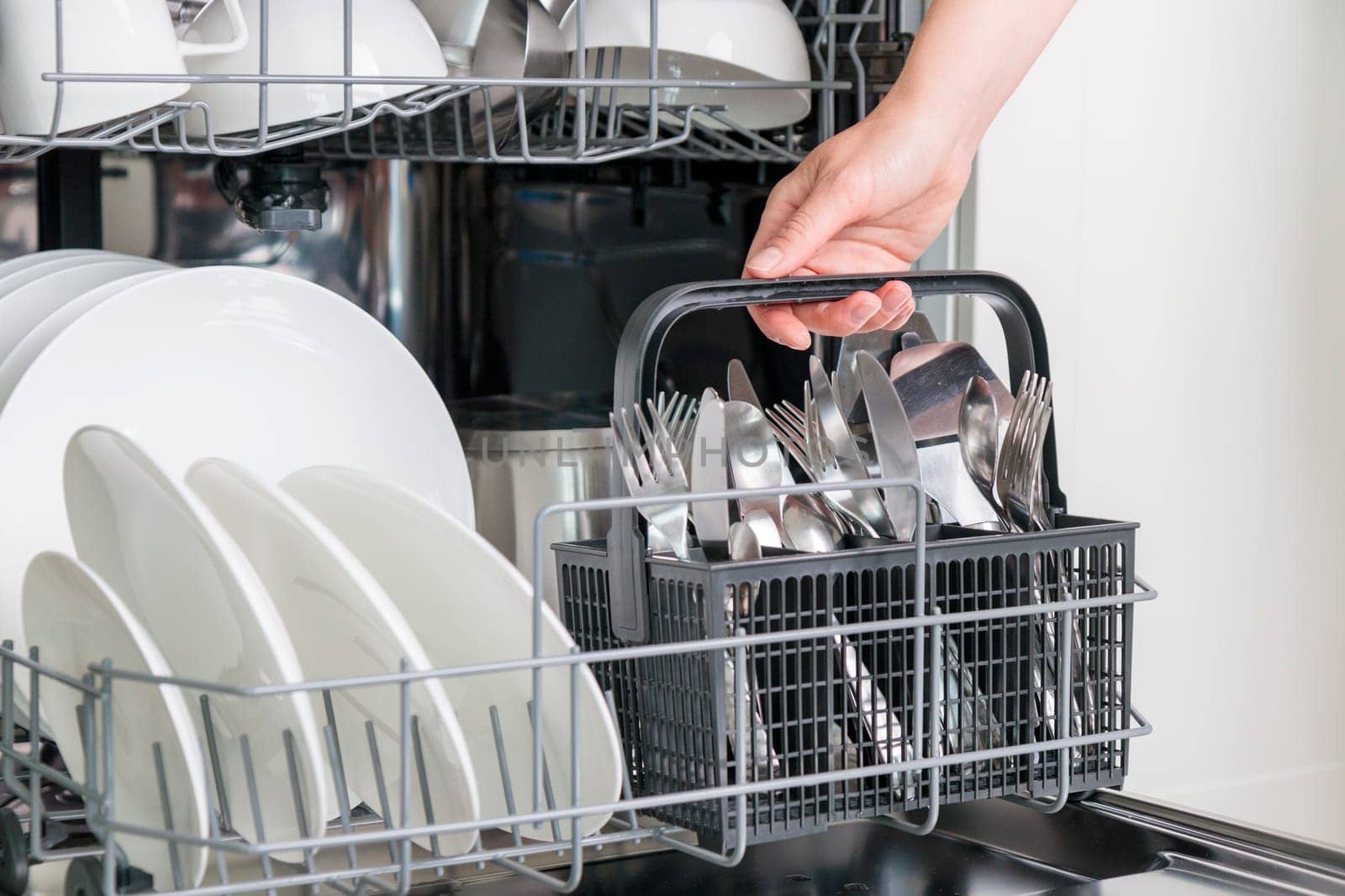 Female hand unloading dishwasher with tableware. Kitchen appliances, lifestyle view. Woman takes a cutlery basket from the dishwasher. Concept of modern technologies in the kitchen