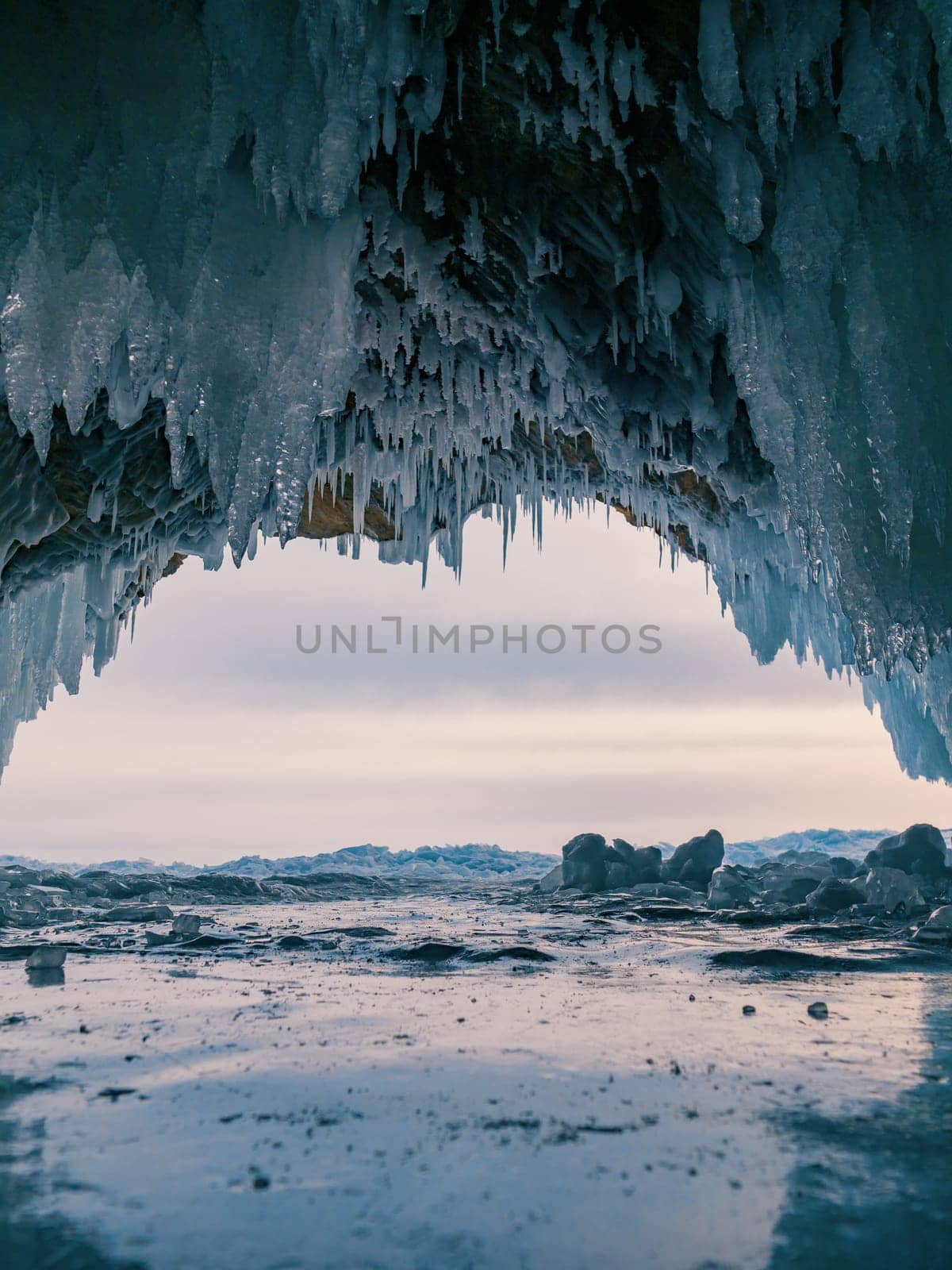 Inside a stunning ice cave on Lake Baikal, large icicles hang from the ceiling, creating a breathtaking winter landscape. Snow-covered mountains can be seen far in the distance. by Busker