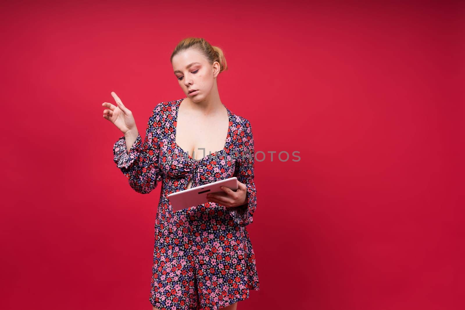 A woman in a purple floral dress is holding a tablet computer, her arm raised in a gesture. She has a microphone pinned to her onepiece garment, with lipstick and flash photography visible